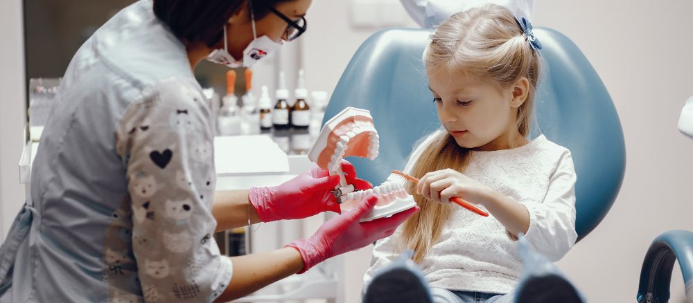 Dentista infantil atendiendo a niña, enseñando cepillado dental. Cuidado dental